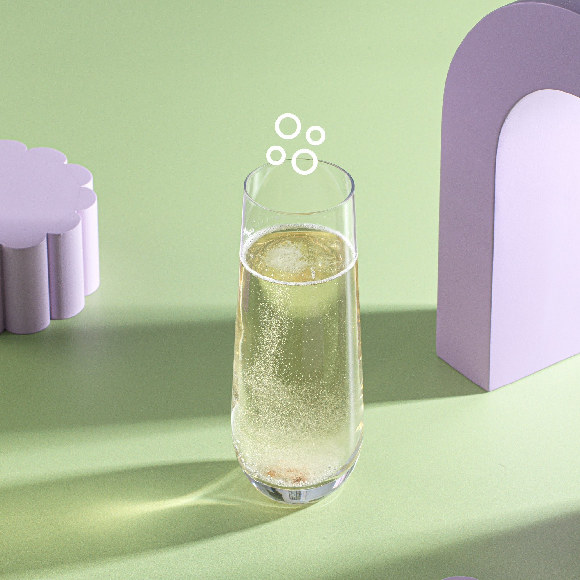 A Milo Champagne Glass on a green surface. There are two abstract shapes, both purple in the background. 