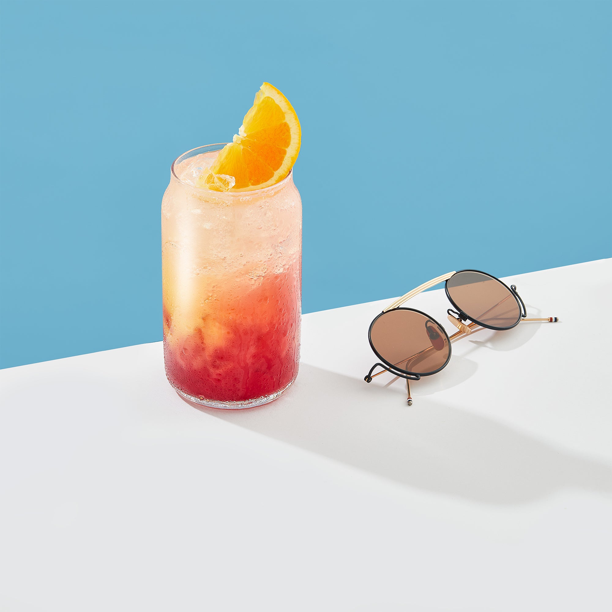 A glass of orange/strawberry juice and sunglasses on a table, next to Classic Can Shape Tumbler Drinking Glass Cups by JoyJolt.