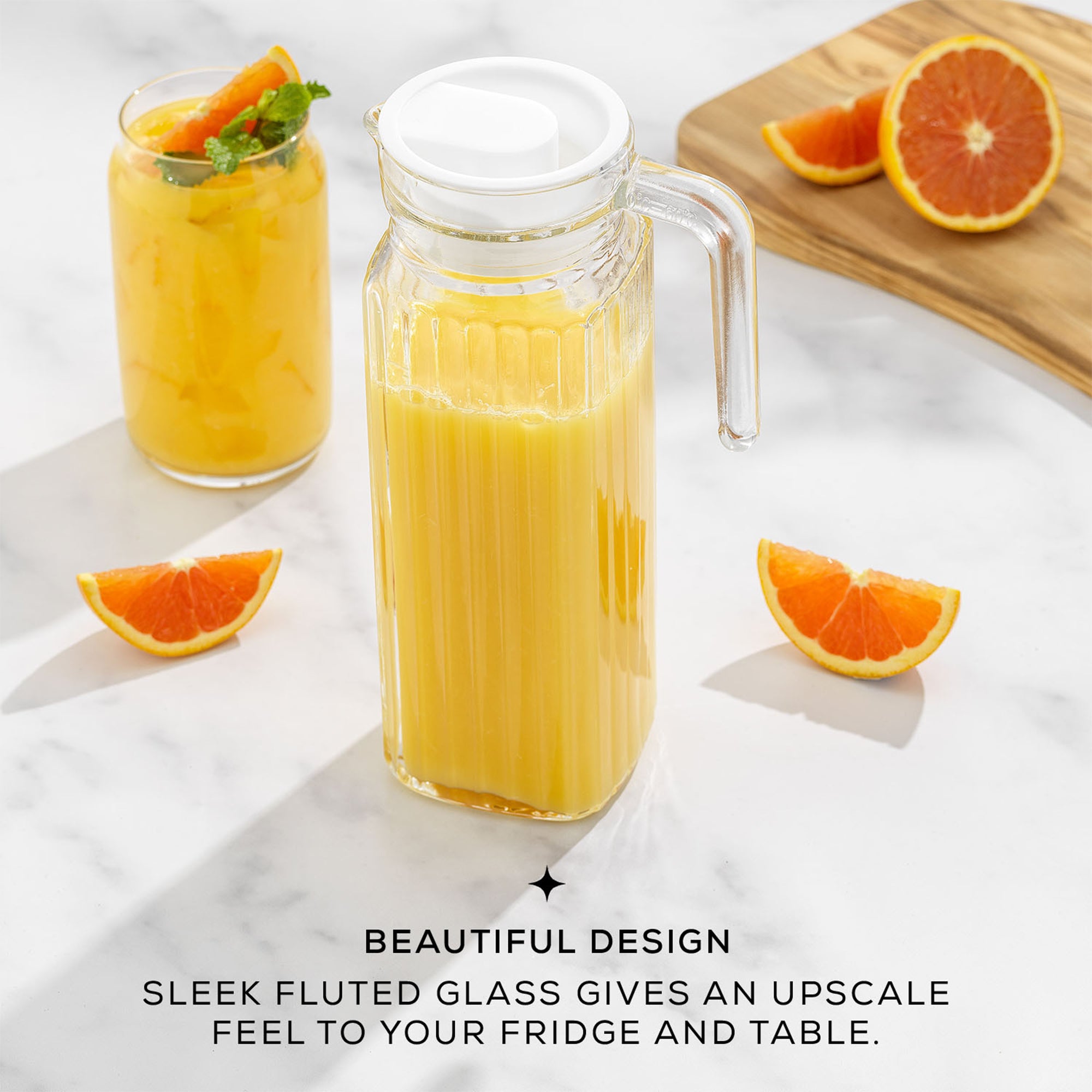 A large, clear glass pitcher filled with orange juice sits on a table next to a pile of oranges. The pitcher has a sleek, fluted design and a lid. Text on the image reads “BEAUTIFUL DESIGN. SLEEK FLUTED GLASS GIVES AN UPSCALE FEEL TO YOUR FRIDGE AND TABLE”.
