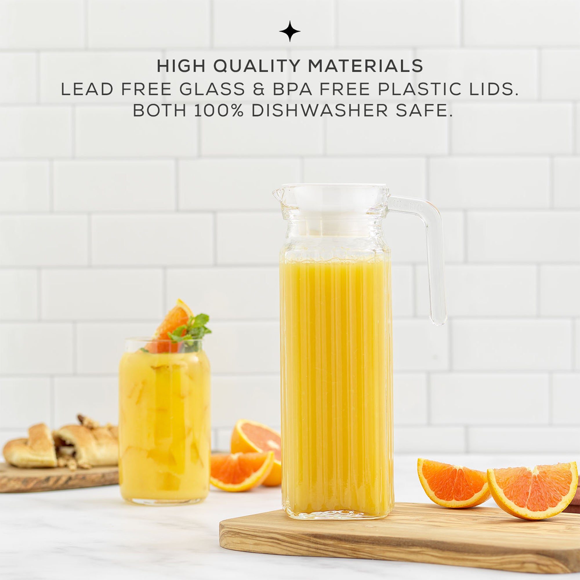 A glass pitcher filled with orange juice sits on a table next to a pile of oranges. The pitcher has a clear glass body, a white lid, and a handle for easy pouring. Text on the image reads "HIGH QUALITY MATERIALS LEAD FREE GLASS & BPA FREE PLASTIC LIDS. BOTH 100% DISHWASHER SAFE." This is a JoyJolt Beverage Serveware Glass Pitcher, 40oz.
