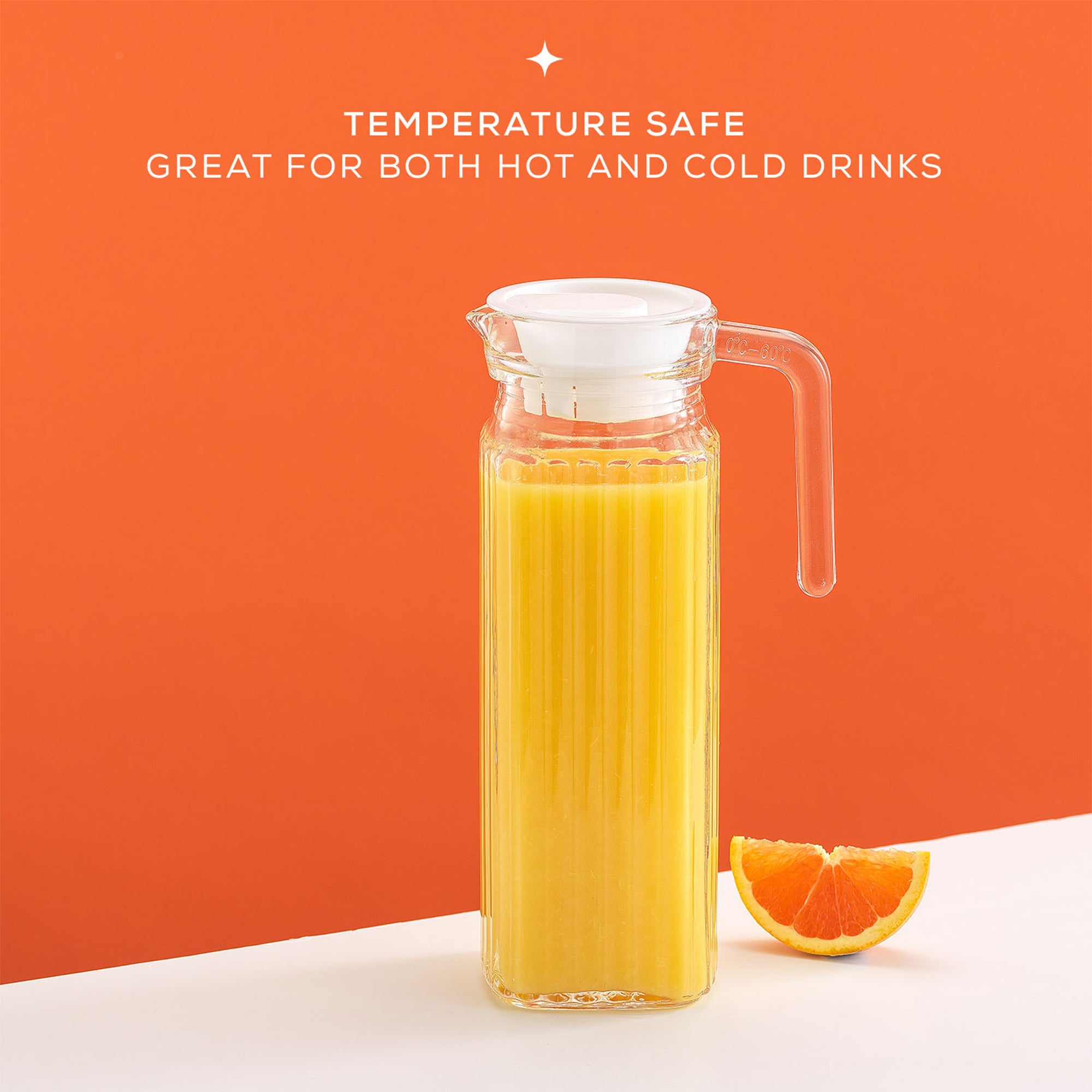 A large, clear glass pitcher filled with orange juice sits on a table next to a single orange. The pitcher has a sleek, fluted design and a lid. Text on the image reads “TEMPERATURE SAFE. GREAT FOR BOTH HOT AND COLD DRINKS”. This is a JoyJolt Beverage Serveware Glass Pitcher, 40oz.