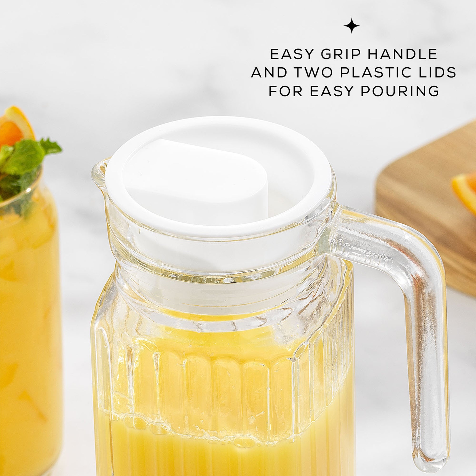 A 40-ounce JoyJolt Beverage Serveware Glass Pitcher filled with orange juice sits on a table. The pitcher has a clear glass body, a white lid, and a handle for easy pouring. Text on the image reads "EASY GRIP HANDLE AND TWO PLASTIC LIDS FOR EASY POURING".