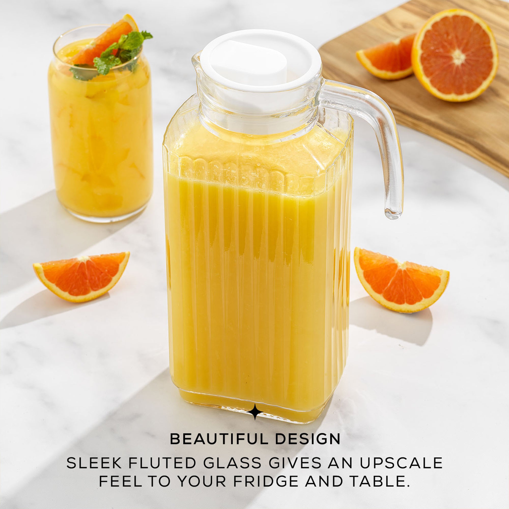 A large, clear glass pitcher filled with orange juice sits on a table next to a pile of oranges. The pitcher has a sleek, fluted design and a lid.  Text on the image reads  “BEAUTIFUL DESIGN. SLEEK FLUTED GLASS GIVES AN UPSCALE BEAUTIFUL DESIGN FEEL TO YOUR FRIDGE AND TABLE.”  This is a JoyJolt Beverage Serveware Glass Pitcher, 60oz.