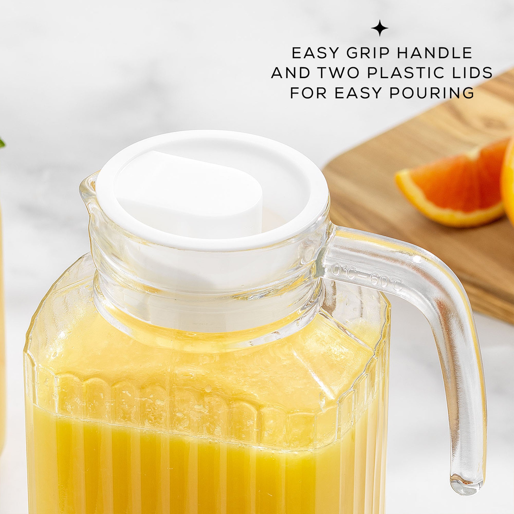 A clear glass pitcher with a handle sits on a table. The pitcher is filled with orange juice and has a white lid. Text on the image reads "EASY GRIP HANDLE AND TWO PLASTIC LIDS FOR EASY POURING". This is a JoyJolt Beverage Serveware Glass Pitcher, 60oz.