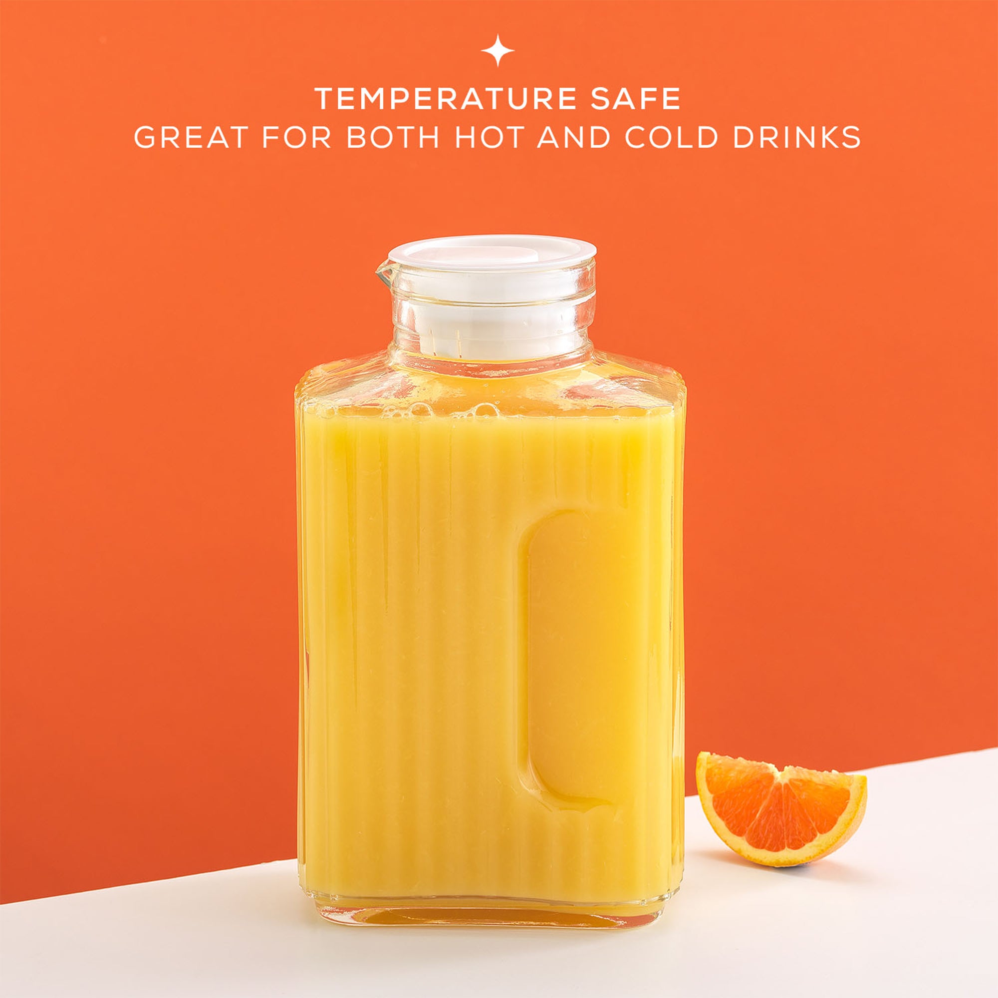 A large, glass JoyJolt Beverage Serveware Glass Pitcher filled with orange juice sits on a table next to a whole orange. The pitcher has a leak-proof lid and a handle for easy pouring. Text on the image reads: "TEMPERATURE SAFE. GREAT FOR BOTH HOT AND COLD DRINKS."