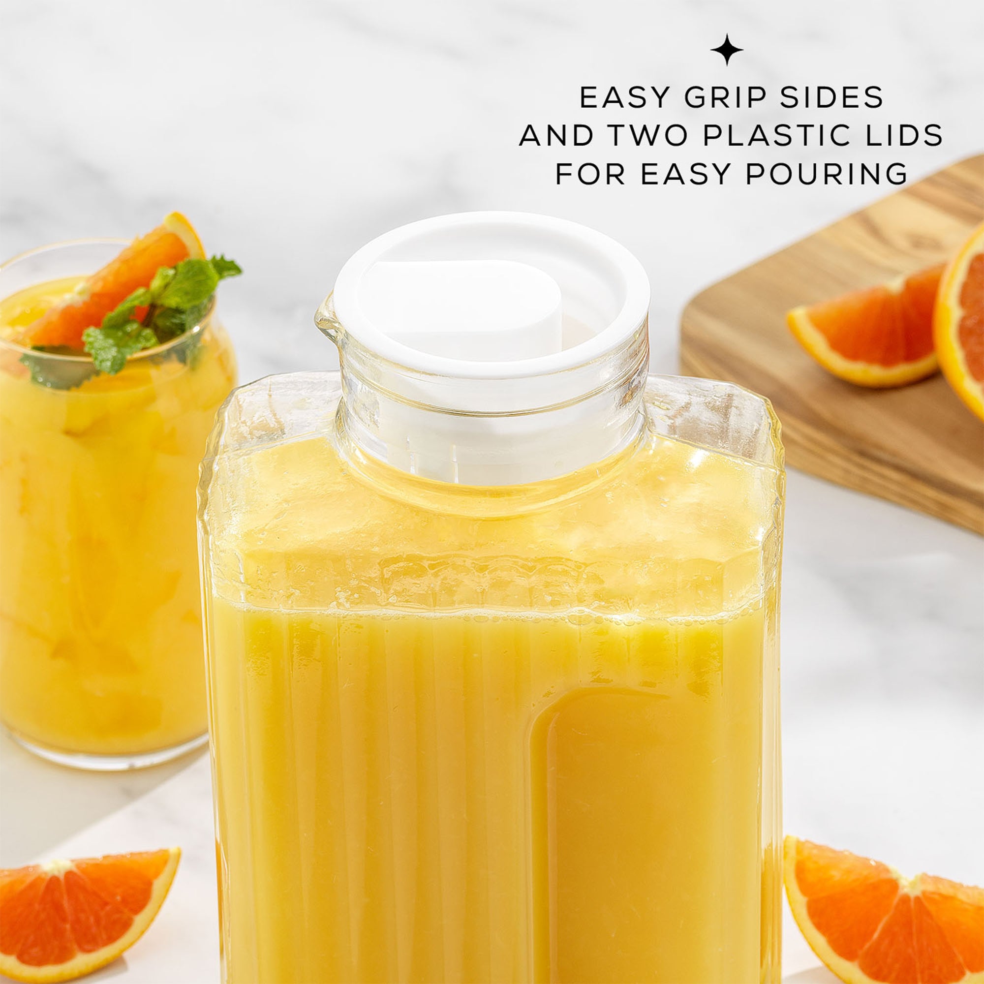 A clear square-shaped JoyJolt Beverage Serveware Glass Pitcher filled with orange juice, featuring easy-grip sides and two plastic lids for simple pouring, with fresh orange slices in the background.