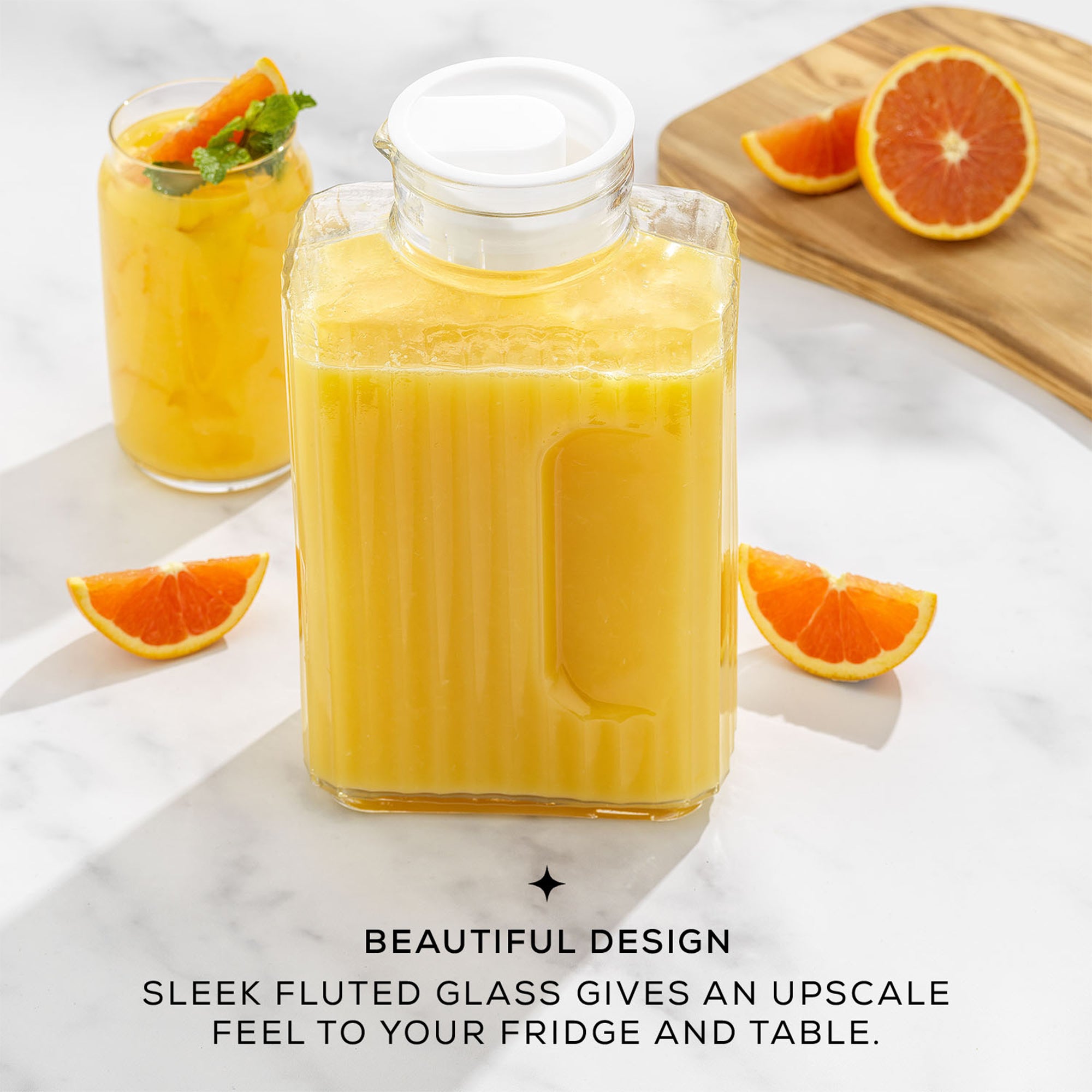 A JoyJolt Beverage Serveware Glass Pitcher filled with orange juice sits on a table next to a cluster of oranges. The 2-liter pitcher has a sleek fluted design. Text on the image reads: "SLEEK FLUTED GLASS GIVES AN UPSCALE BEAUTIFUL DESIGN FEEL TO YOUR FRIDGE AND TABLE."