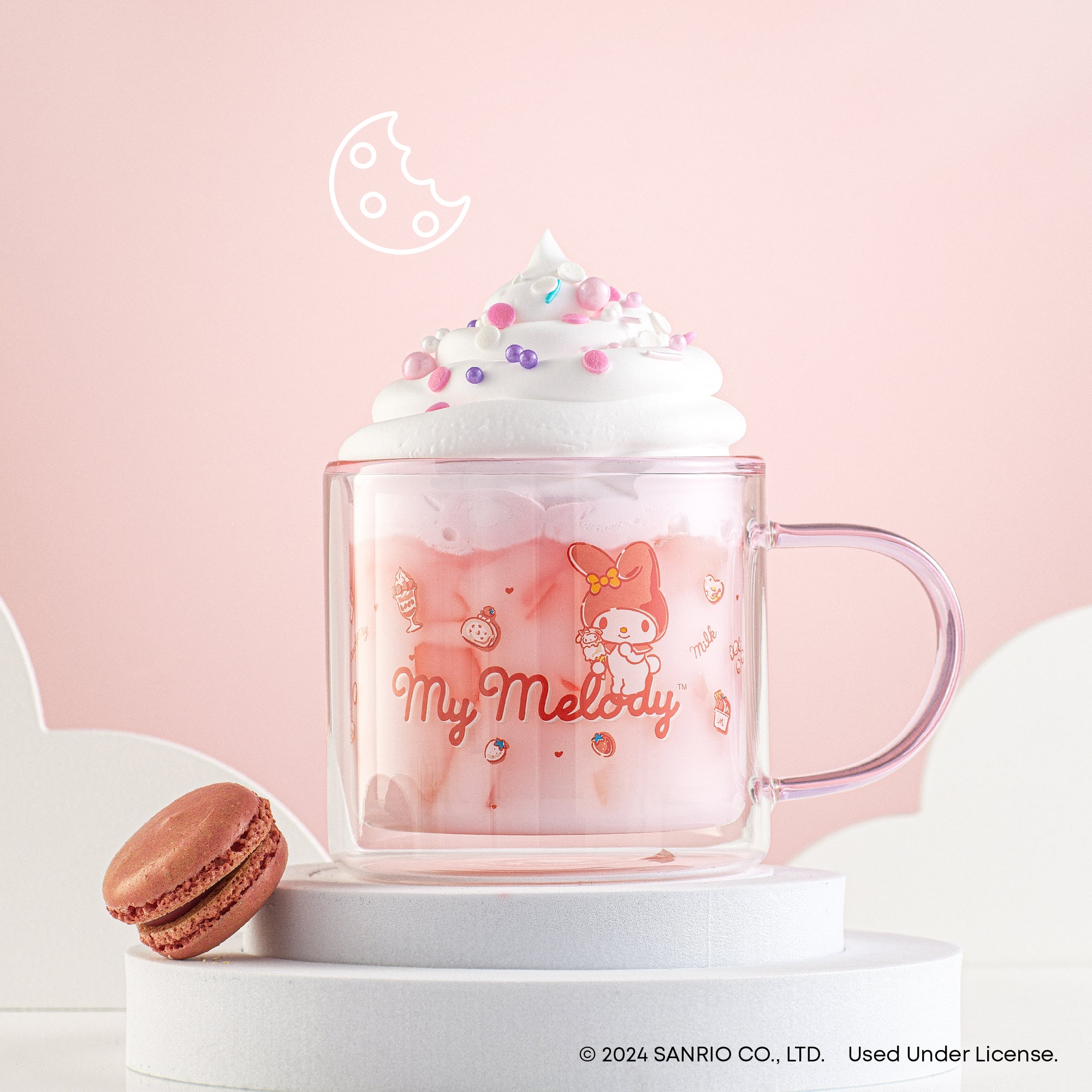 A pink JoyJolt My Melody mug filled with a creamy beverage. Next to the mug is a chocolate macaron. The mug has an image of My Melody by Sanrio. 