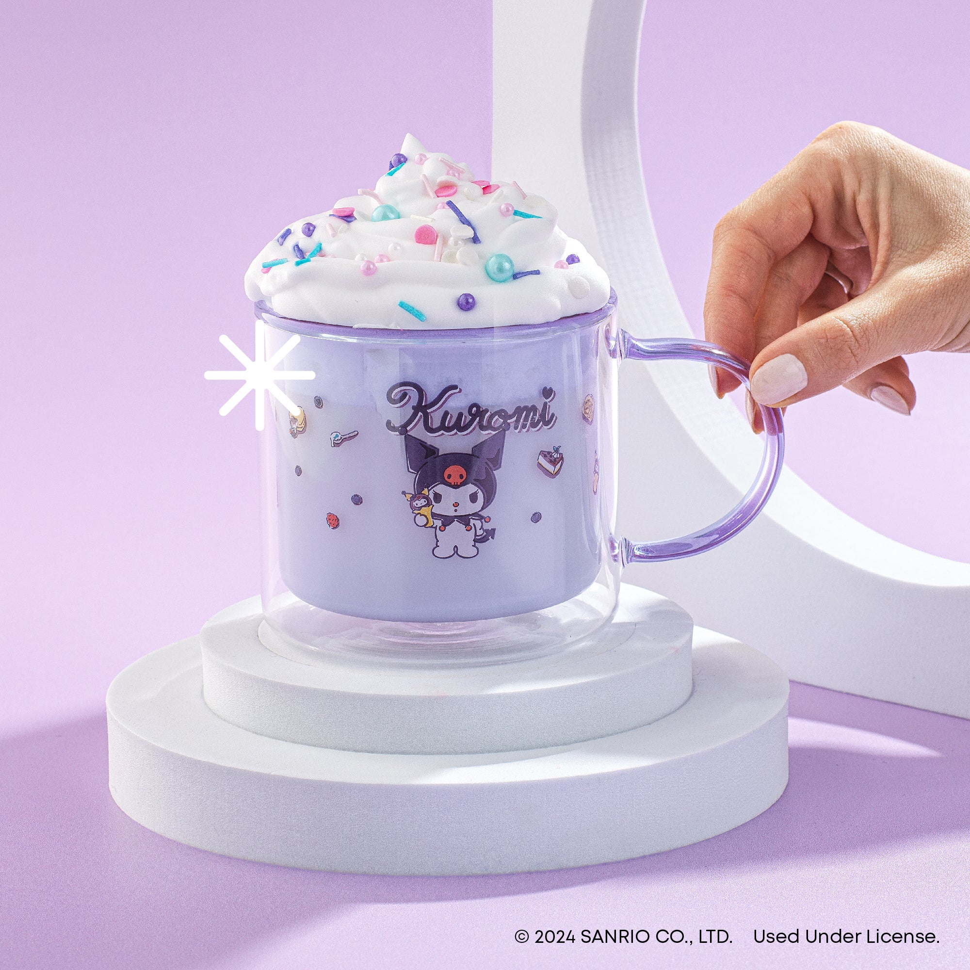 A person holding a clear glass mug with whipped cream and sprinkles on top. The mug has a purple Kuromi logo and the text "Kuromi".