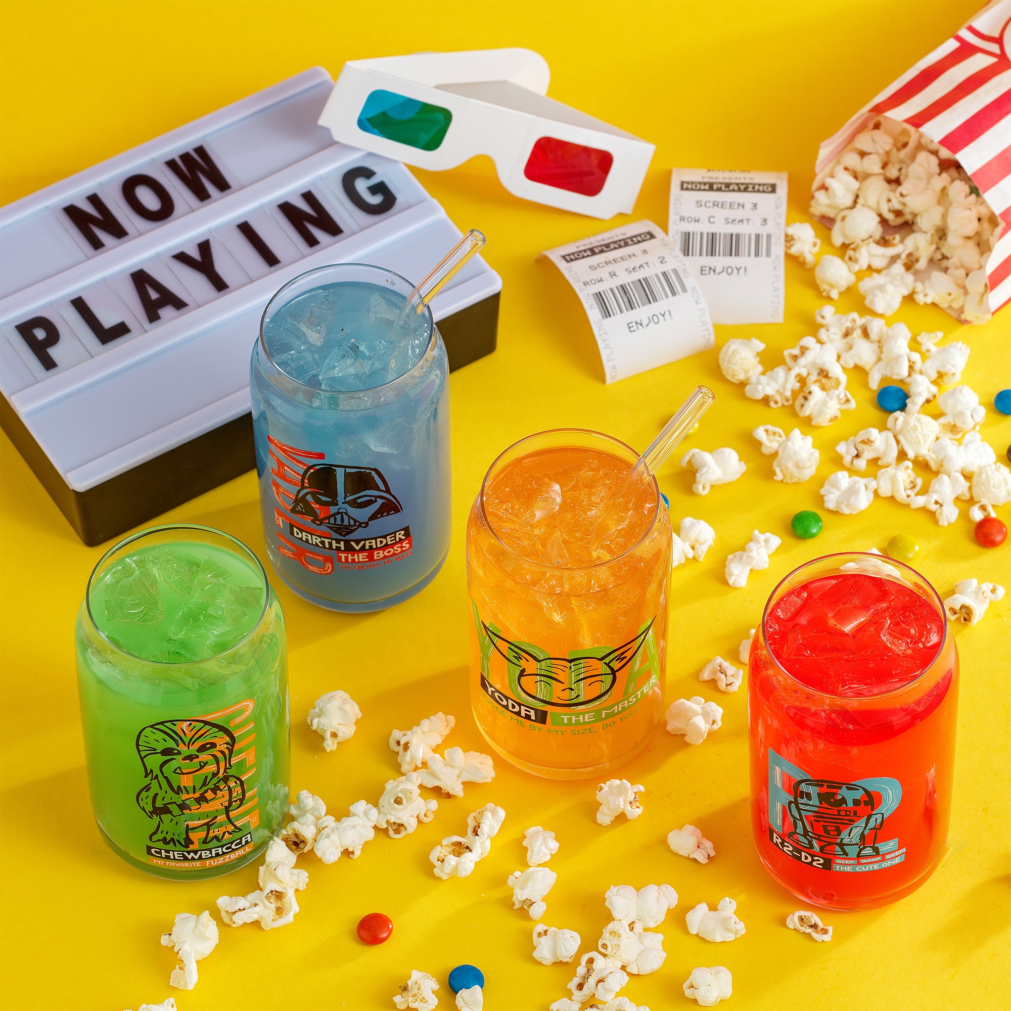 JoyJolt's Star Wars 'Now Playing' glass tumbler set featuring Yoda, Chewbacca, Darth Vader, R2-D2. They are all surrounded by popcorn, candied chocolate, ticket stubs, and 3D glasses. A 'Now Playing' movie sign is placed alongside it. 
