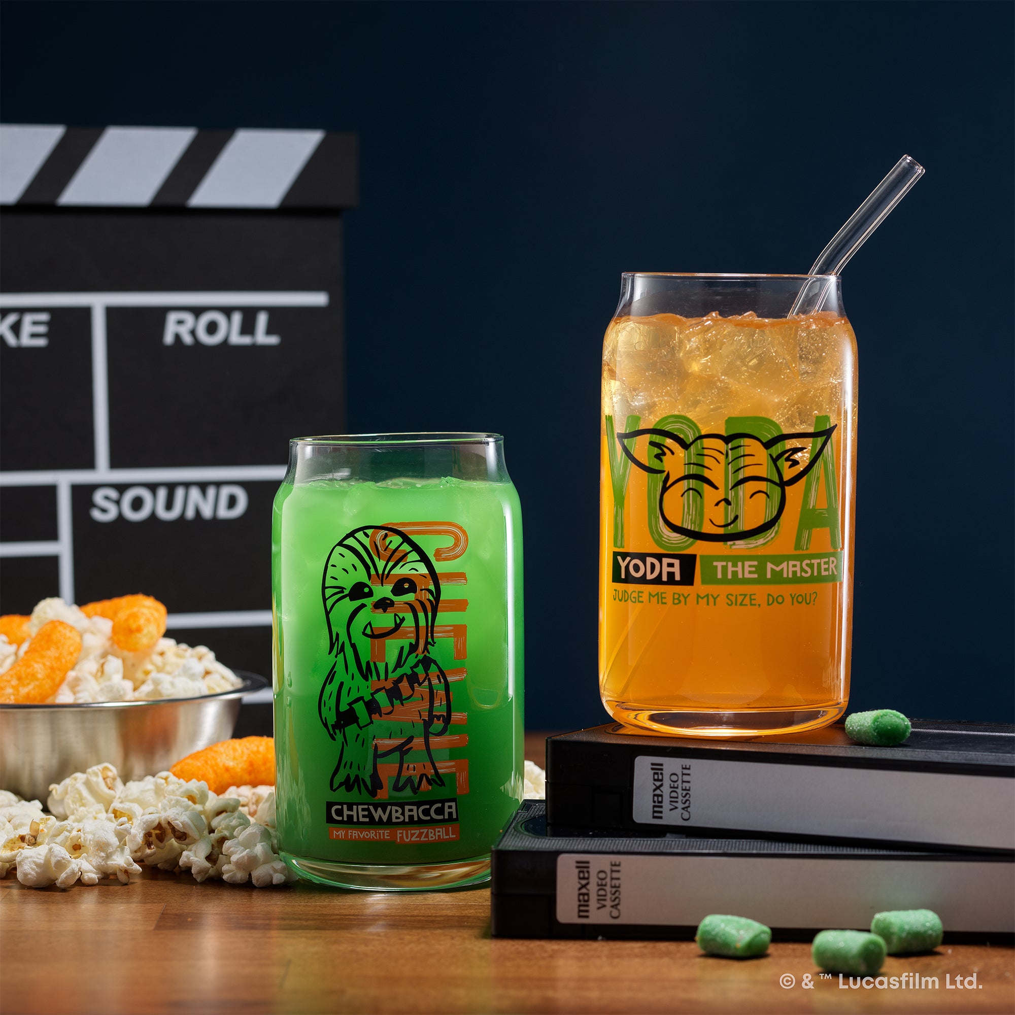 JoyJolt's Star Wars 'Now Playing' glass tumbler set featuring Chewbacca and Yoda. Yoda's glass is on two VHS tapes with a bowl of popcorn and chips in the background. 